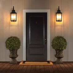 two urns with plants are on the front porch next to a black door and light fixture