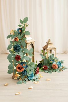 an arrangement of flowers and greenery on the floor next to two small gold lanterns