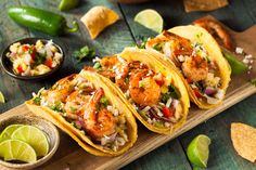 three shrimp tacos on a cutting board with limes, salsa and tortillas