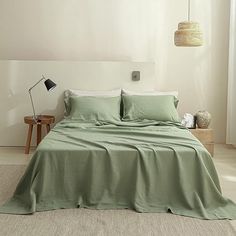 a bed with green sheets and pillows in a white room next to a lamp on the wall