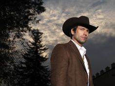 a man wearing a brown suit and cowboy hat standing in front of a cloudy sky