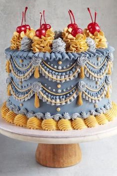 a blue and yellow decorated cake with cherries on top, sitting on a pedestal