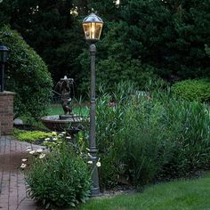 a lamp post sitting in the middle of a garden next to a brick walkway and water fountain