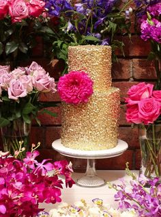 a three tiered cake with pink flowers and greenery in front of a brick wall