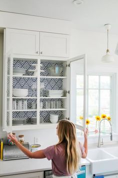 a woman standing in a kitchen next to a sink and cabinets with sunflowers on the window sill