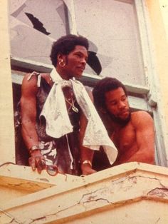 two black men standing on the ledge of a window sill looking out at bats