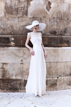 a woman in a white dress and hat leaning against a wall with her hands on her hips