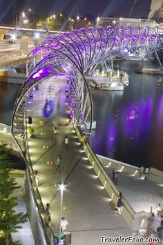 people are walking on the walkway next to the water at night with purple lights in the background