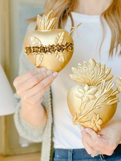 a woman holding two golden vases with flowers on them, one in the shape of an apple