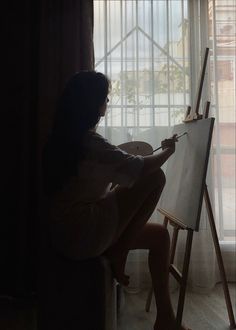 a woman sitting in front of a window drawing on an easel next to a window