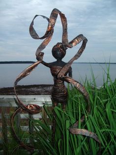 a metal sculpture sitting in the grass next to some tall green grass with water in the background