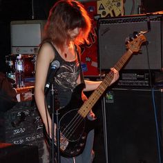 a woman standing in front of a microphone and holding a bass guitar with her right hand