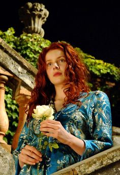 a woman with red hair is holding a white rose in her hand and looking off into the distance