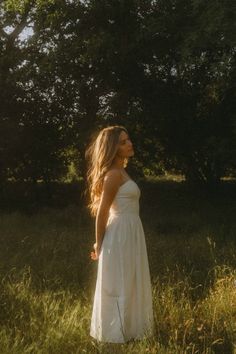 a woman in a white dress standing in tall grass with trees in the back ground