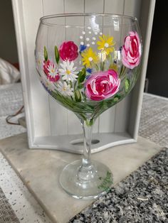 a painted wine glass sitting on top of a counter next to a white box with flowers in it