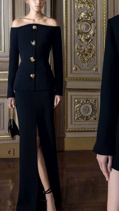 a woman standing in front of a wall wearing a black suit with gold buttons on it