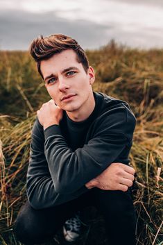 a young man sitting in the grass with his arms crossed and looking at the camera