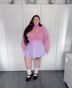 a woman with long hair wearing a pink sweater and purple skirt standing in front of white cabinets