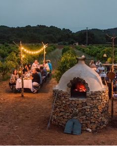 people sitting around an outdoor pizza oven with lights strung from the roof and on poles