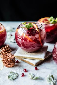 three glasses filled with red liquid and garnished with orange slices, cranberries, pine cones and mint