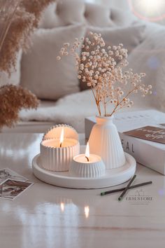 two white candles sitting on top of a table next to a book and some flowers