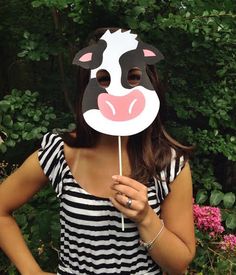 a woman wearing a cow mask holding a stick in front of her face with trees and bushes behind her