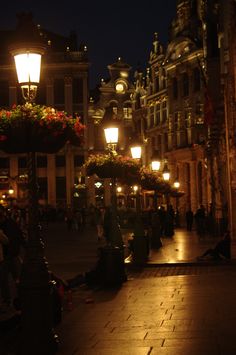 people are sitting on the sidewalk in front of some buildings at night with street lights