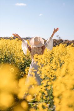 a woman standing in a yellow field with her arms spread out to the side, wearing a white dress and hat