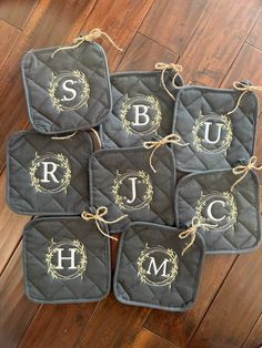 four black pot holders with gold monogrammed letters and wreaths on them, sitting on a wood floor