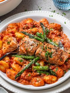 a white bowl filled with pasta and meat covered in tomato sauce, asparagus and potatoes