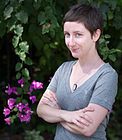 a woman with her arms crossed standing in front of some purple flowers and greenery