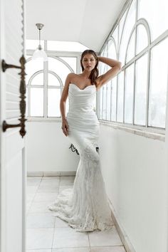 a woman in a white dress is standing by a window with her hand on her head