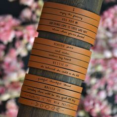 leather bracelets with words written on them sitting on a wooden pole in front of pink flowers