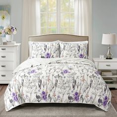 a white bed with purple flowers on it and a window in the backround