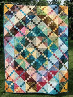 a colorful quilt hanging from a clothesline on the grass in front of some trees