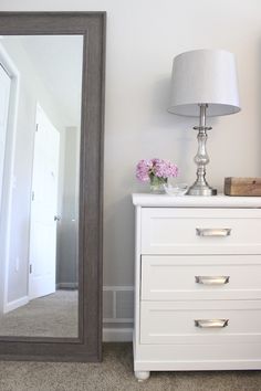 a white dresser sitting next to a large mirror on top of a wooden floor under a lamp