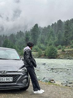 a man standing next to a black car near a river and forest in the background