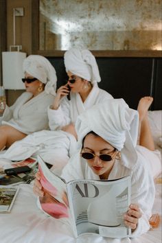 three women in bathrobes are sitting on a bed and one is reading a magazine