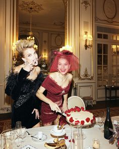 two women in red dresses cutting into a strawberry covered cake on top of a table