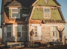 two men are standing in front of an old house with green shingles and white windows