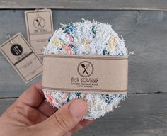 a hand holding a ball of yarn with two tags attached to it and another item in the background