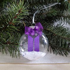 a glass ornament with a purple bow hanging from a christmas tree branch and snow flakes