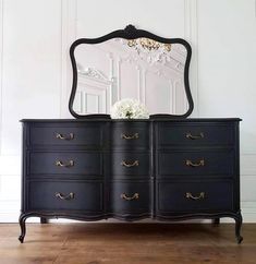 a black dresser with an ornate mirror on top and flowers in the bottom, against a white wall