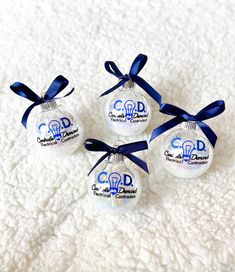 four glass ornaments with blue ribbon tied around them on a white blanketed surface, each ornament shaped like a light bulb