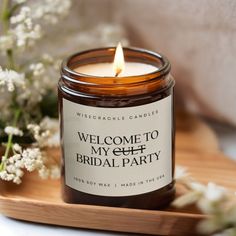 a candle that is sitting on a tray with some flowers in the background and welcome to my self bridal party sign