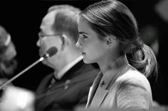 two people sitting at a table with microphones in front of them and one person standing behind the woman