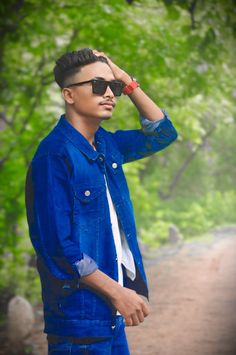 a young man wearing sunglasses standing on a dirt road in front of some green trees