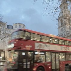 a red double decker bus driving down a street next to tall buildings in the background