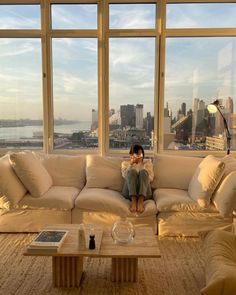 a woman sitting on top of a couch in front of a window next to a coffee table