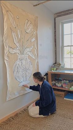 a woman sitting on the floor in front of a wall hanging with a flower vase